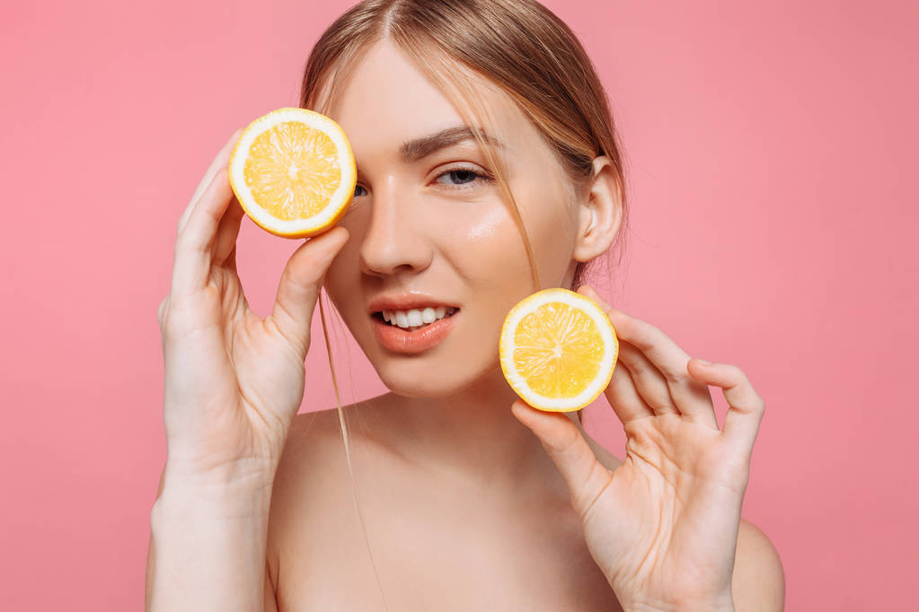 Portrait of a cheerful feminine girl, natural clear skin, a girl with two pieces of lemon, covering one eye with a lemon, isolated on a pink background. Skin care concept, natural beauty - Photo, Image