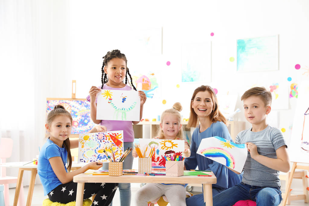 Söpöt pienet lapset opettajan kanssa näyttämässä maalauksiaan sisätiloissa
 - Valokuva, kuva