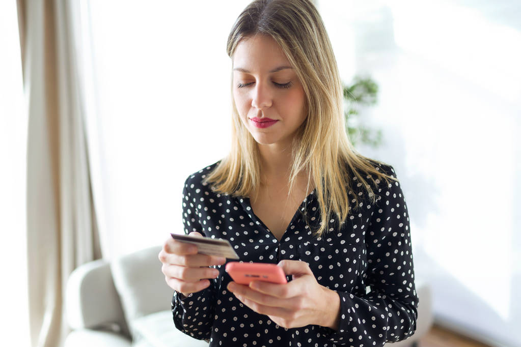 Prise de vue de jeune femme concentrée tenant une carte de crédit blanche pour faire du shopping en ligne avec téléphone portable dans le salon à la maison
. - Photo, image
