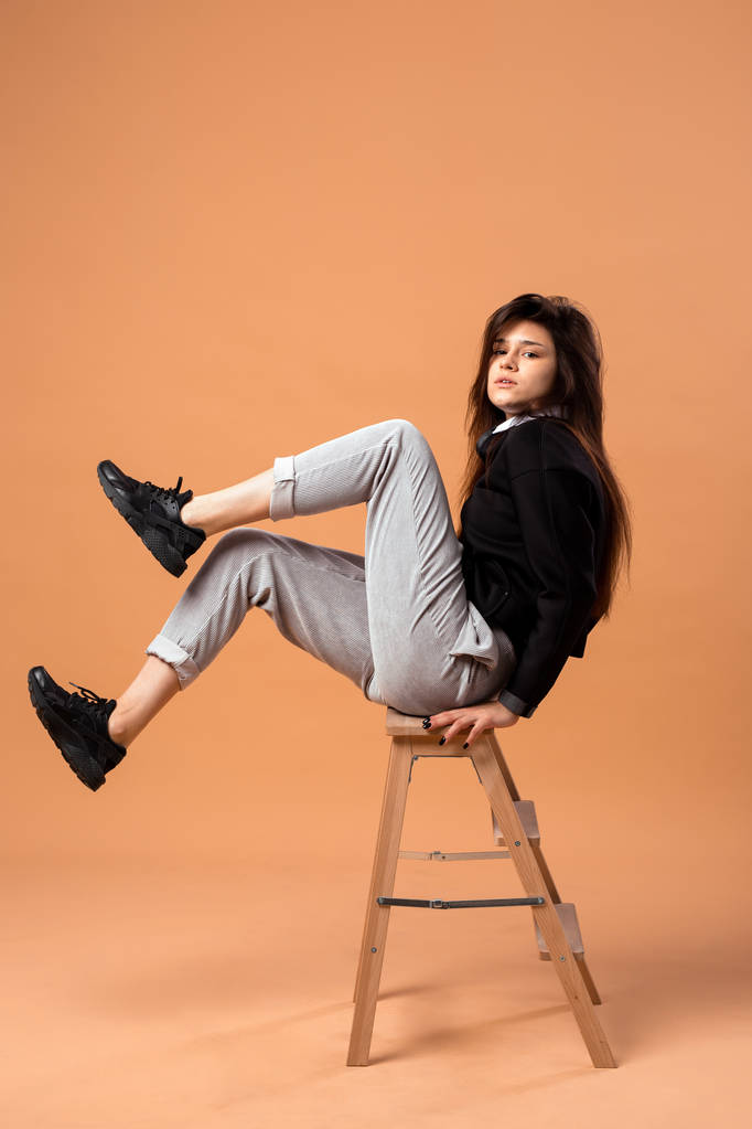 Chica de pelo oscuro vestida con camisa blanca, pantalones grises, chaqueta negra y zapatillas negras está sentada en un taburete de madera en el fondo beige en el estudio
 - Foto, Imagen