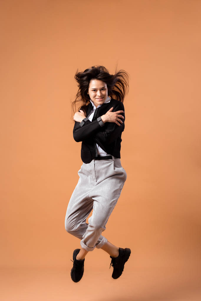 Happy dark-haired girl dressed in white shirt, gray trousers, black jacket  and black sneakers jumps on the beige background in the studio - Photo, Image