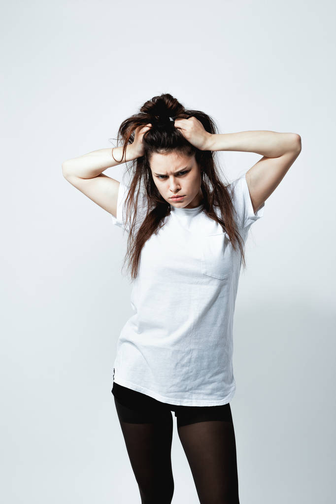 Stylish young dark-haired girl with a funny hairstyle dressed in white t-shirt, black tights and shoes is posing on the white background in the studio - Photo, Image