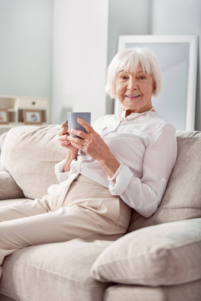 Charmante Seniorin posiert mit Tasse Tee auf Couch - Foto, Bild