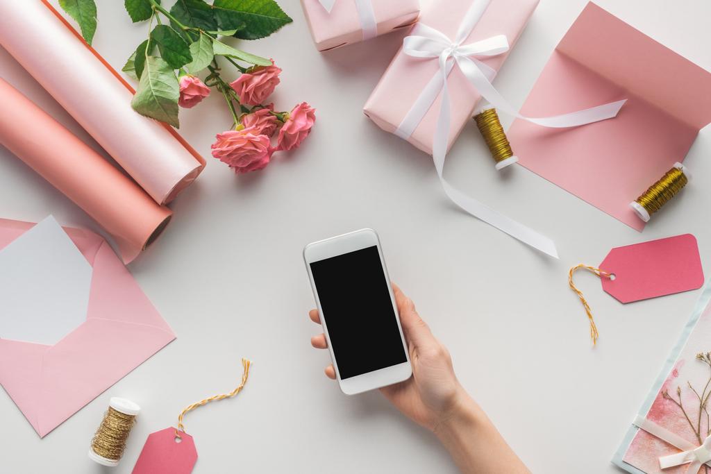 vista recortada de la mujer sosteniendo teléfono inteligente cerca de rosas, rollos de papel, regalos envueltos, sobre, tarjeta de felicitación y carretes de hilo sobre fondo gris
   - Foto, imagen