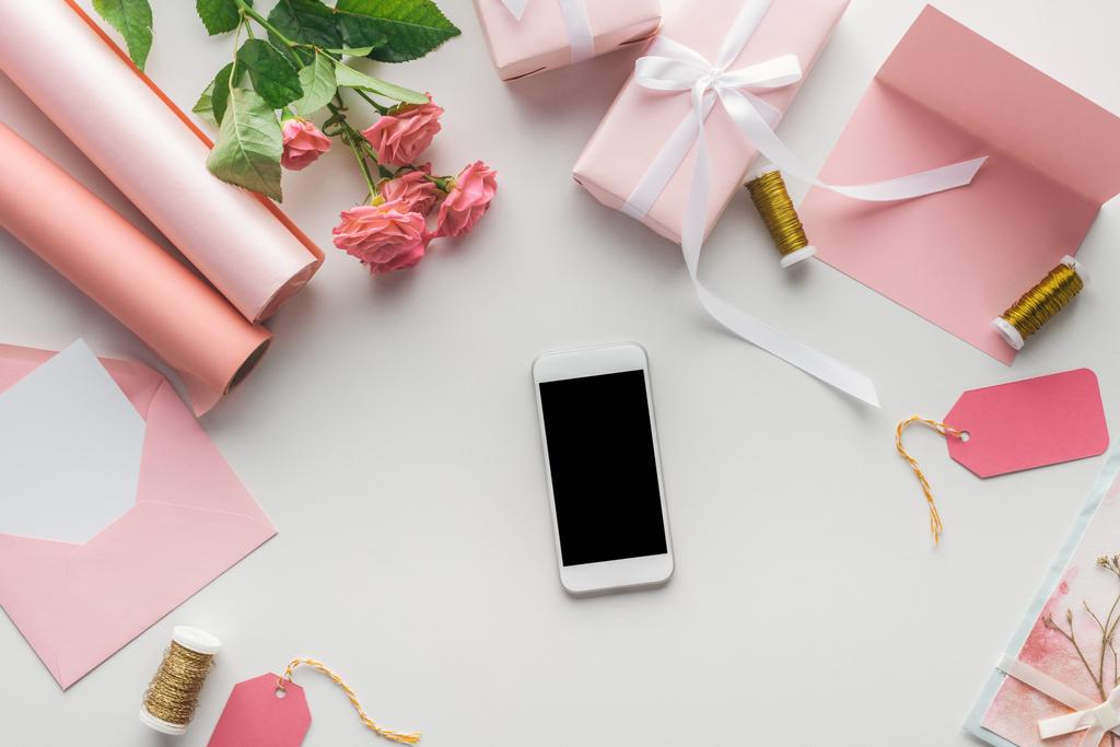 Ansicht des Smartphones in der Nähe von Rosen, Papierrollen, verpackten Geschenken, Umschlägen und Garnspulen auf grauem Hintergrund   - Foto, Bild
