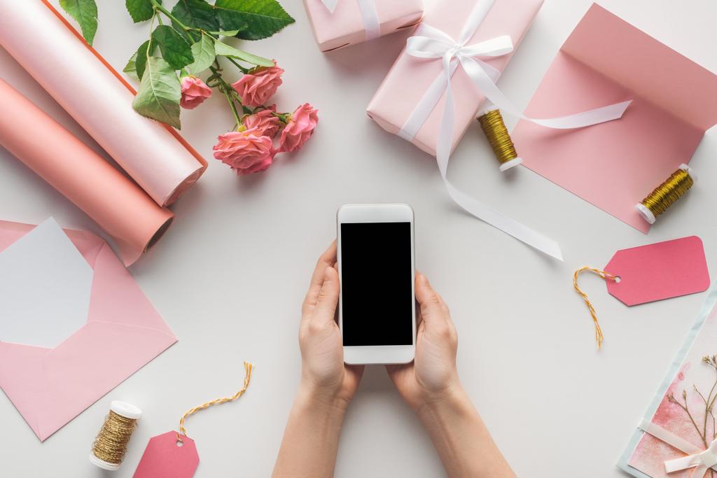 Ausgeschnittene Ansicht einer Frau mit Smartphone in der Nähe von Rosen, Papierrollen, verpackten Geschenken, Umschlag, Karte und Fadenspulen auf grauem Hintergrund - Foto, Bild
