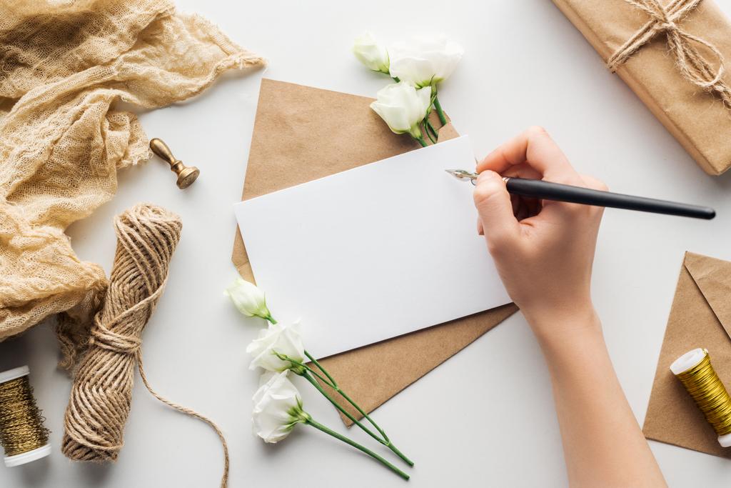 bijgesneden beeld van vrouw schrijven in kaart met inkt pen in de buurt van eustoma, doek, envelop en verpakt geschenk op grijze achtergrond  - Foto, afbeelding