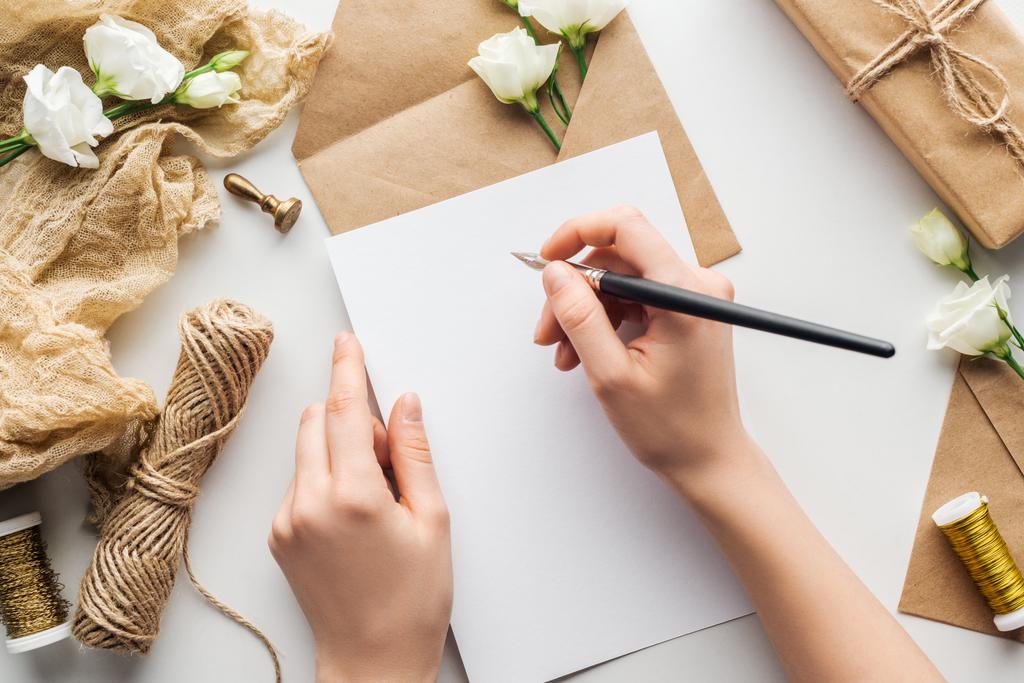 bijgesneden beeld van vrouw schrijven in kaart met inkt pen in de buurt van bloemen, doek, envelop en verpakt geschenk op grijze achtergrond  - Foto, afbeelding