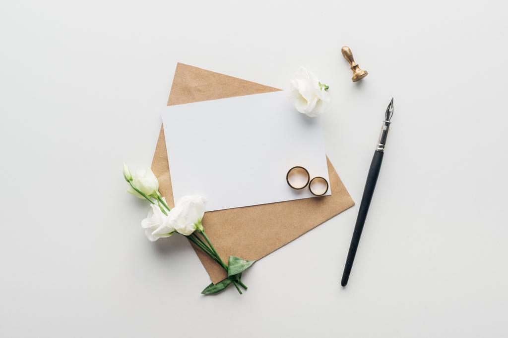 vista superior del sobre con tarjeta vacía, pluma de tinta, flores, sello y anillos de boda dorados sobre fondo gris
 - Foto, Imagen