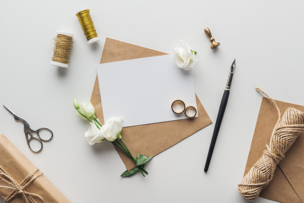 灰色の背景の空のカード、ボールペン、花、スタンプ、ギフト包装、はさみ、スレッドと黄金の結婚指輪のスプールのエンベロープのトップ ビュー - 写真・画像