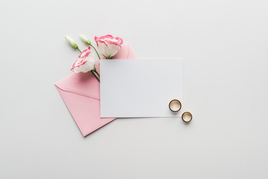 pohled shora na prázdnou kartu s růžovou obálku, květiny a zlaté snubní prsteny na šedém pozadí - Fotografie, Obrázek