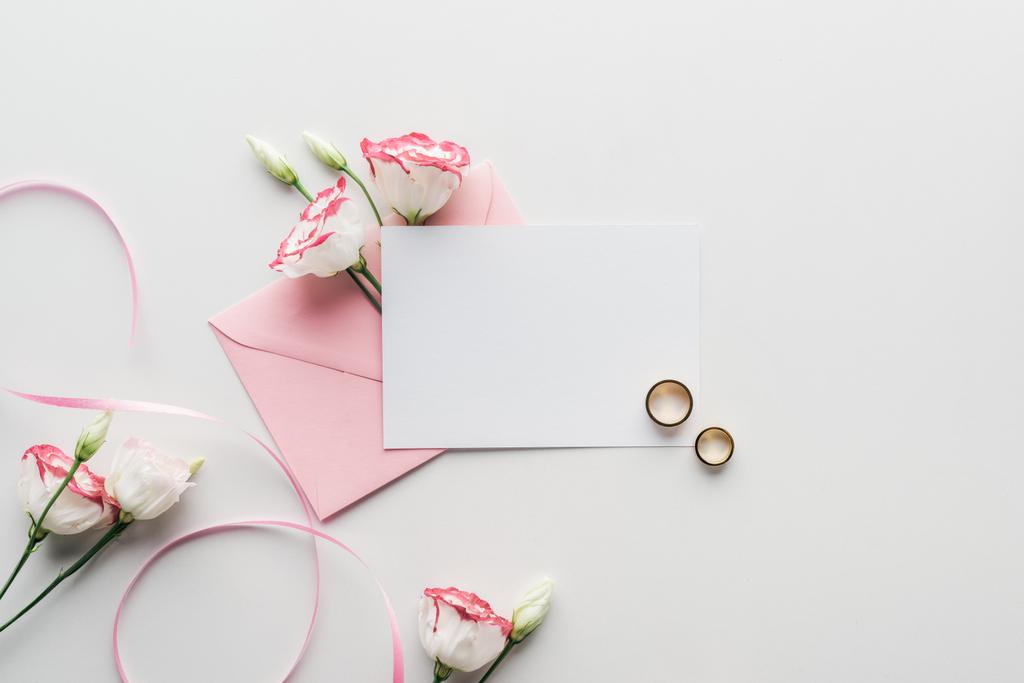 вид сверху на пустую открытку с розовым конвертом, цветами, шелковой лентой и золотыми обручальными кольцами на сером фоне
 - Фото, изображение