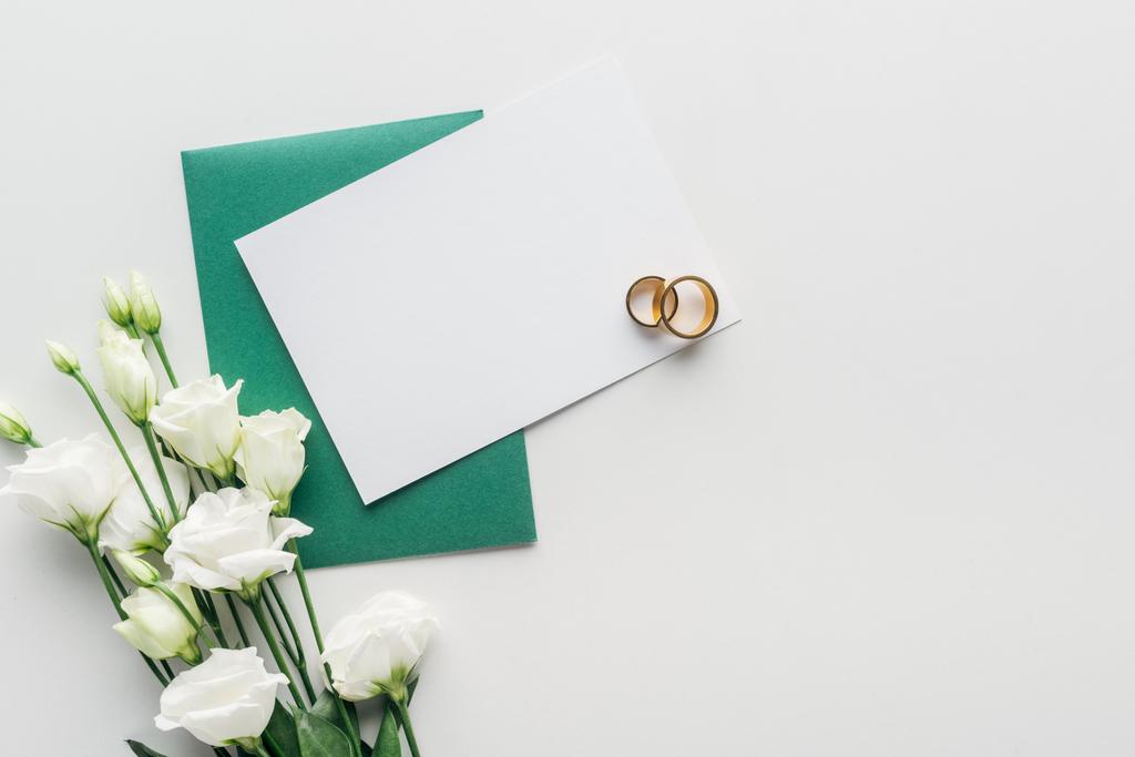 top view of empty card with green envelope, flowers and golden wedding rings on grey background - Photo, Image