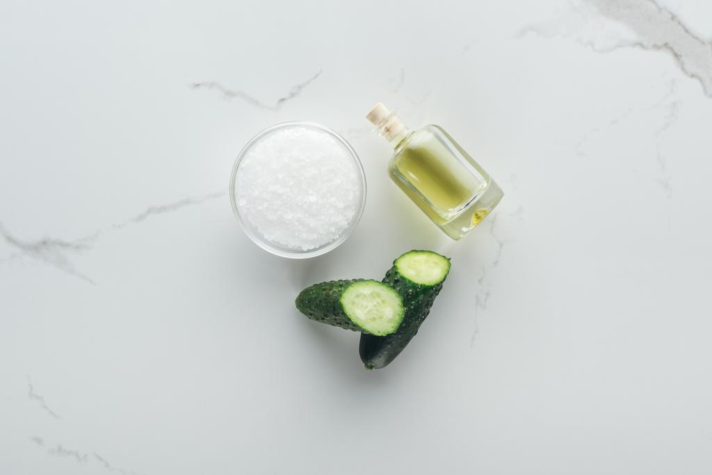 bovenaanzicht van een kom met zout, helften van komkommer en fles komkommer sap op witte ondergrond - Foto, afbeelding