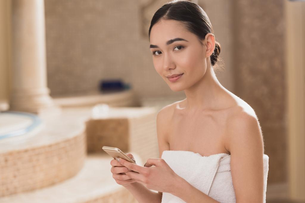 atrakcyjna kobieta azjatyckich w ręcznik za pomocą smartfona i patrząc na kamery  - Zdjęcie, obraz