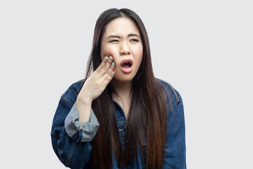 malade belle brune asiatique jeune femme en denim bleu décontracté veste avec maquillage ayant des maux de dents graves et en appuyant sur les doigts à la joue sur fond gris clair
. - Photo, image