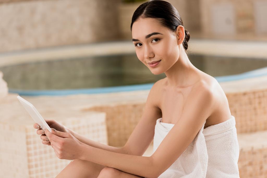 attraktive asiatische Frau im Handtuch mit digitalem Tablet und Blick auf die Kamera  - Foto, Bild