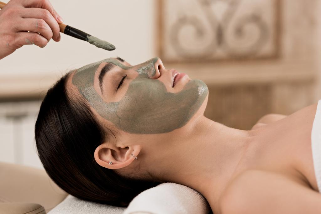 vista lateral de esteticista aplicando máscara de arcilla en la cara de la mujer
 - Foto, Imagen