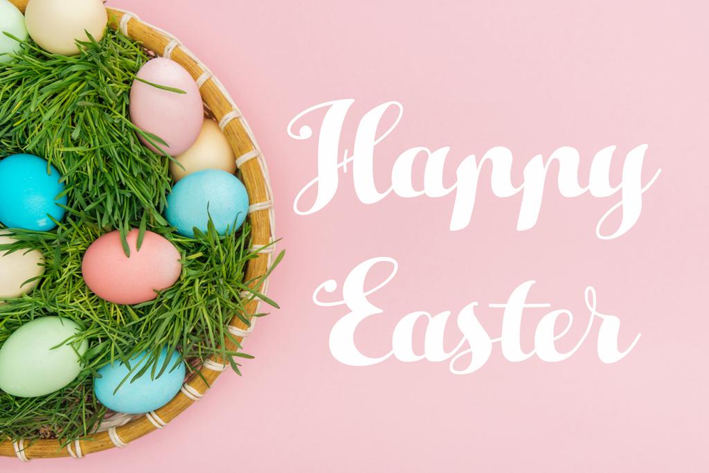 top view of painted eggs in wicker plate with grass isolated on pink with Happy easter lettering  - Photo, Image