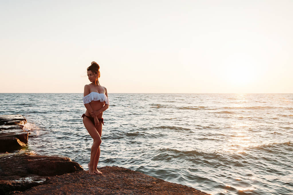 Όμορφη νεαρή γυναίκα που ποζάρει στην παραλία μπροστά στο ηλιοβασίλεμα - Φωτογραφία, εικόνα