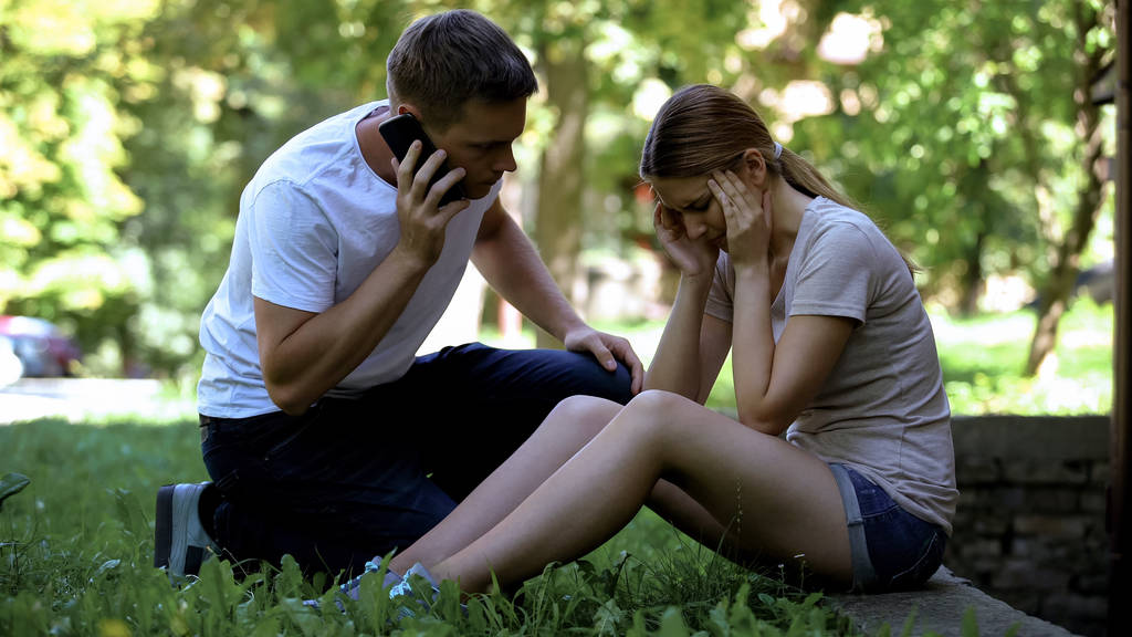 Девушка страдает мигренью, сидит в парке, мужчина вызывает скорую, первая помощь
 - Фото, изображение
