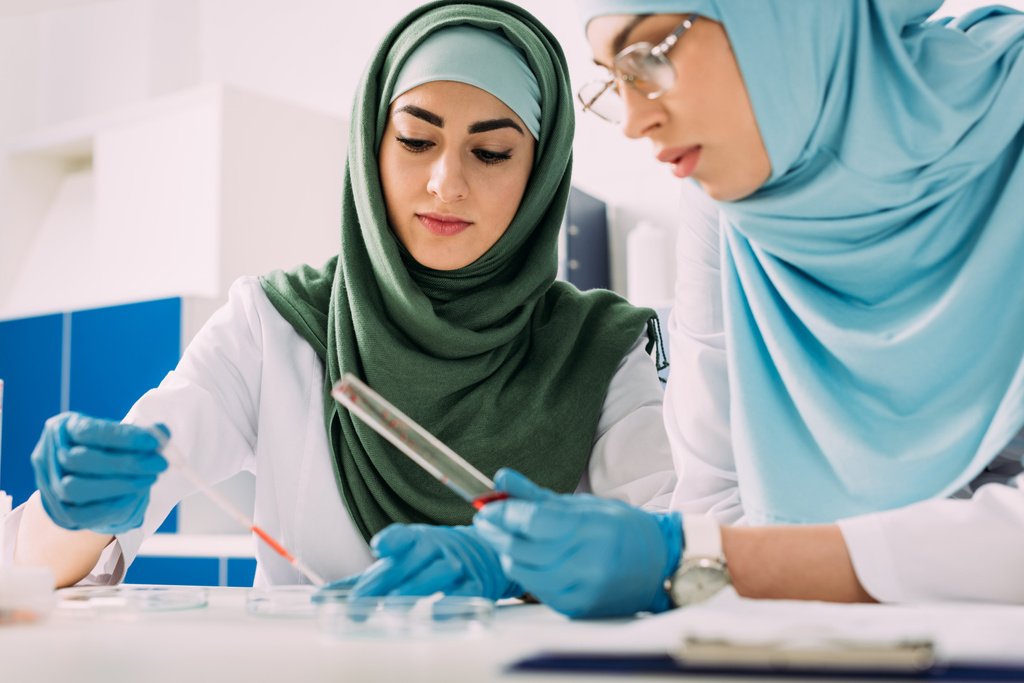 fokussierte muslimische Wissenschaftlerinnen mit Pipette und gläsernem Reagenzglas während eines Experiments im Chemielabor - Foto, Bild
