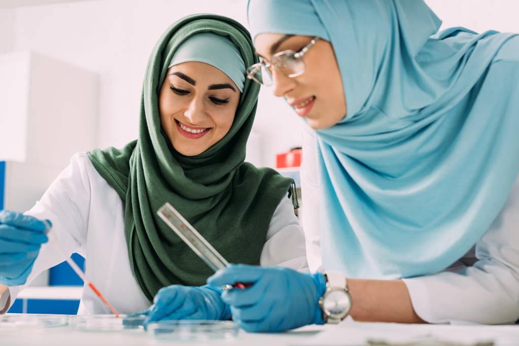 científicos musulmanes sonrientes sosteniendo pipeta y tubo de ensayo de vidrio mientras experimentan en laboratorio químico
 - Foto, Imagen