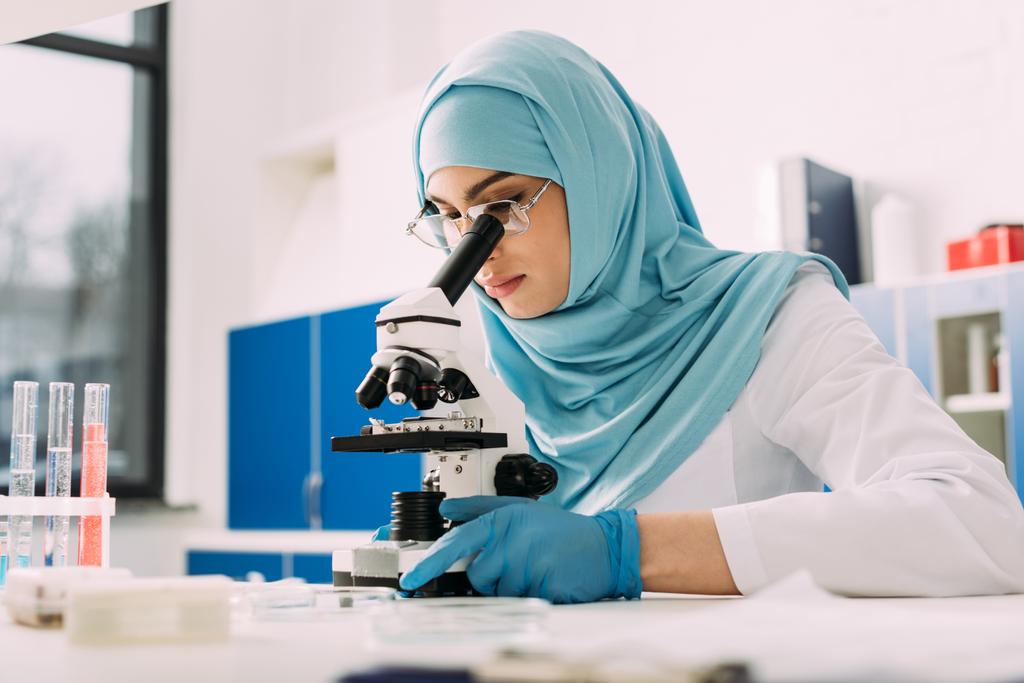συμπυκνωμένο γυναίκα μουσουλμάνος επιστήμονας αναζητούν μέσα από το μικροσκόπιο κατά τη διάρκεια του πειράματος σε χημικό εργαστήριο - Φωτογραφία, εικόνα