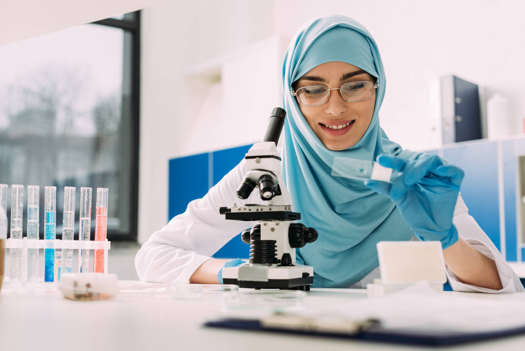 女性イスラム教徒の科学顕微鏡でテーブルに座って、化学実験室での実験中にガラスのサンプルを見て笑みを浮かべてください。 - 写真・画像