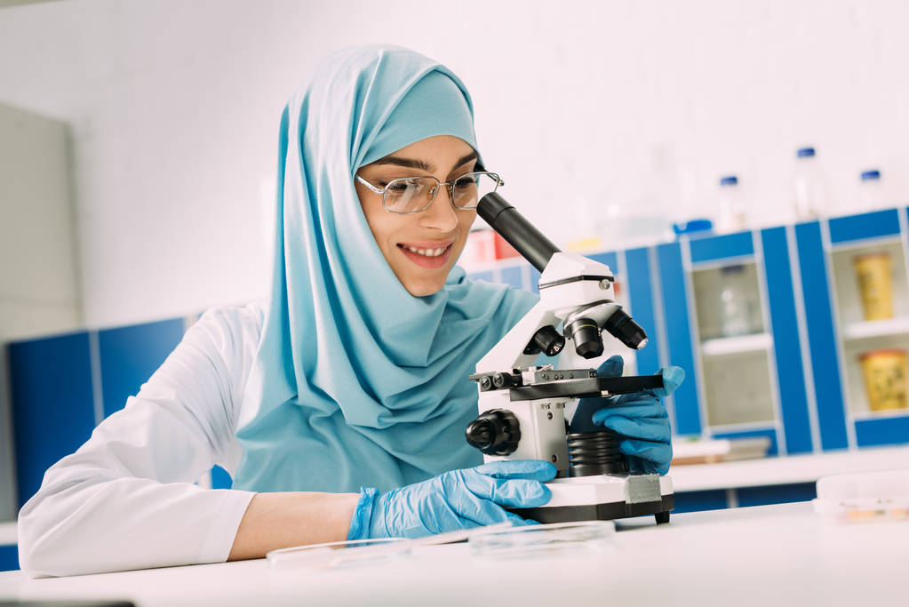 cientista muçulmano sorridente olhando através do microscópio durante o experimento em laboratório químico
 - Foto, Imagem