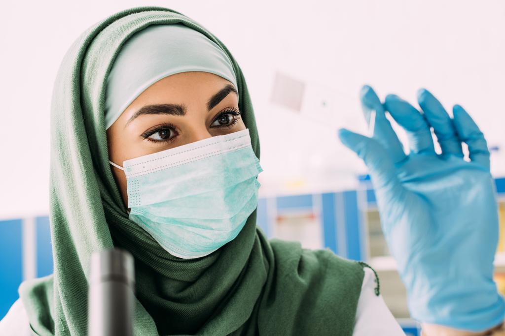 γυναίκα μουσουλμάνος επιστήμονας στην ιατρική μάσκα και χιτζάμπ κρατώντας γυαλί δείγμα κατά τη διάρκεια πειράματος στο εργαστήριο - Φωτογραφία, εικόνα