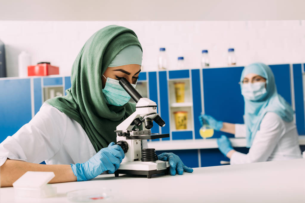 mikroskop ile kimyasal laboratuvar arka planda çalıştığı meslektaşı ile deneme sırasında arayan kadın Müslüman bilim adamı - Fotoğraf, Görsel