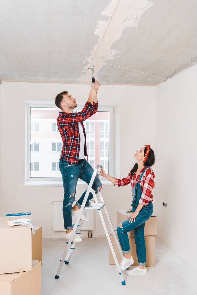 handsome man standing on ladder and changing light bulb near attractive girlfriend - Photo, Image