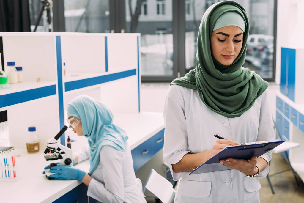 mulheres cientistas muçulmanas concentradas usando microscópio e escrita em prancheta durante experimento em laboratório químico
 - Foto, Imagem