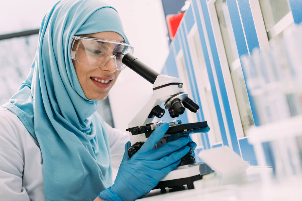 化学研究室での実験中に顕微鏡を通して見る笑顔の女性イスラム教徒の科学者 - 写真・画像