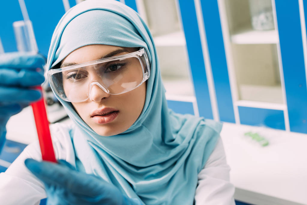 женщина-мусульманка в очках держит пробирку с красной жидкостью в лаборатории
 - Фото, изображение