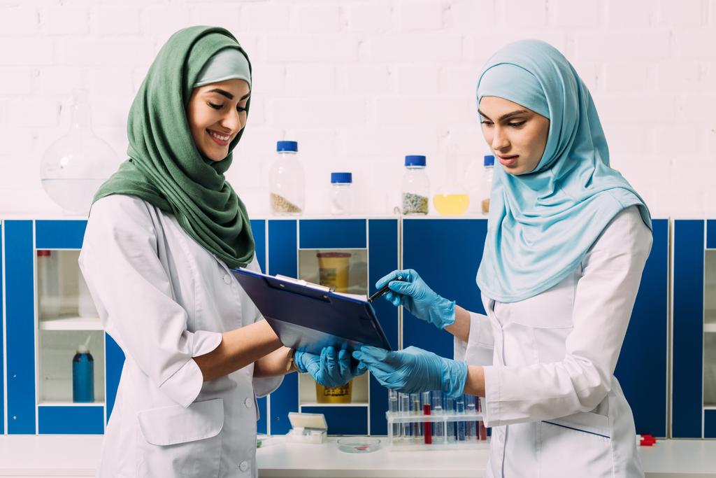 θηλυκό μουσουλμάνων επιστημόνων στο χιτζάμπ κρατώντας πρόχειρο κατά τη διάρκεια του πειράματος σε χημικό εργαστήριο - Φωτογραφία, εικόνα