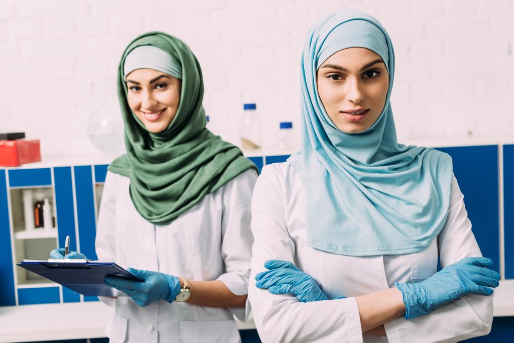 kaunis nainen muslimi kemistit hijabissa kädet ristissä ja leikepöydällä katsot kameraa laboratoriossa
 - Valokuva, kuva