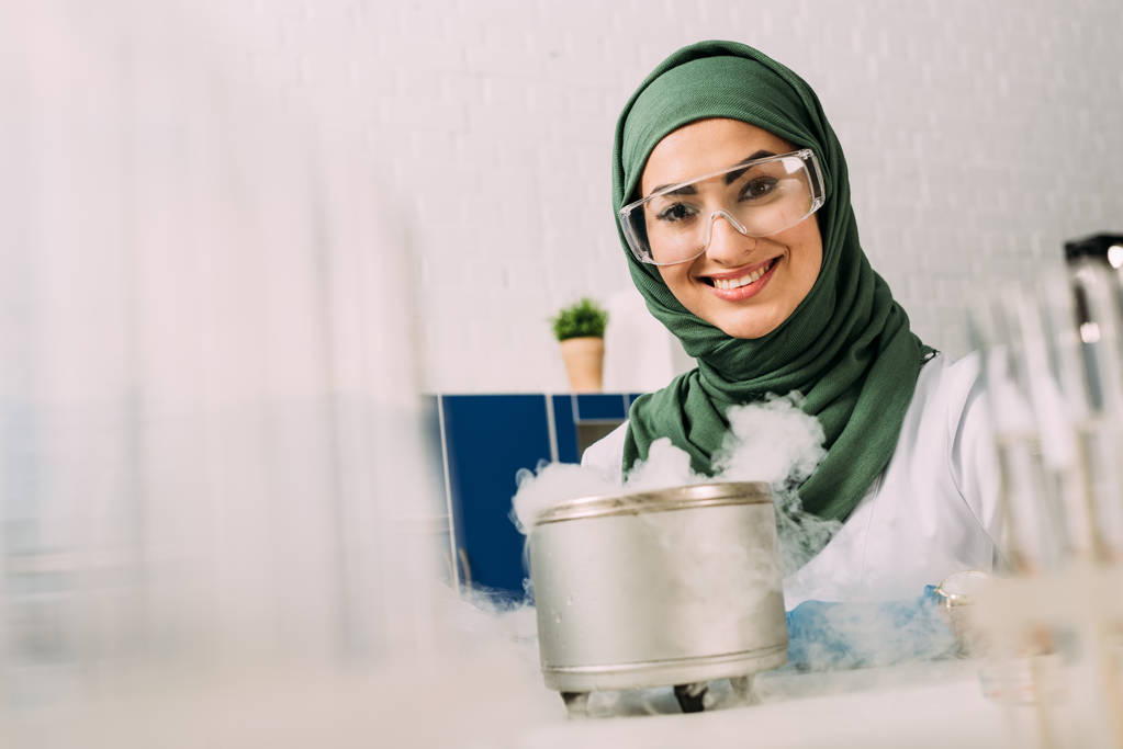 Lächelnde muslimische Wissenschaftlerin blickt während eines Experiments mit Trockeneis im Labor in die Kamera - Foto, Bild