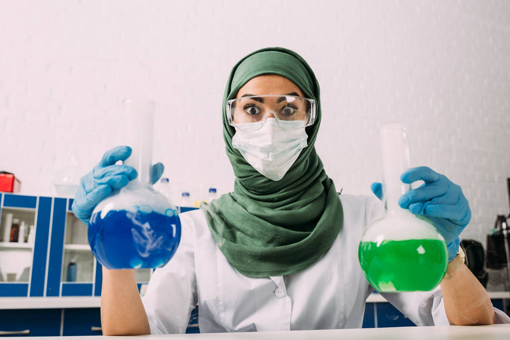 sorprendida científica musulmana mirando a la cámara y sosteniendo frascos durante el experimento en laboratorio químico
 - Foto, imagen