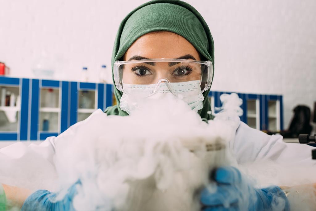 científica musulmana femenina mirando a la cámara mientras experimenta con hielo seco en laboratorio
 - Foto, imagen