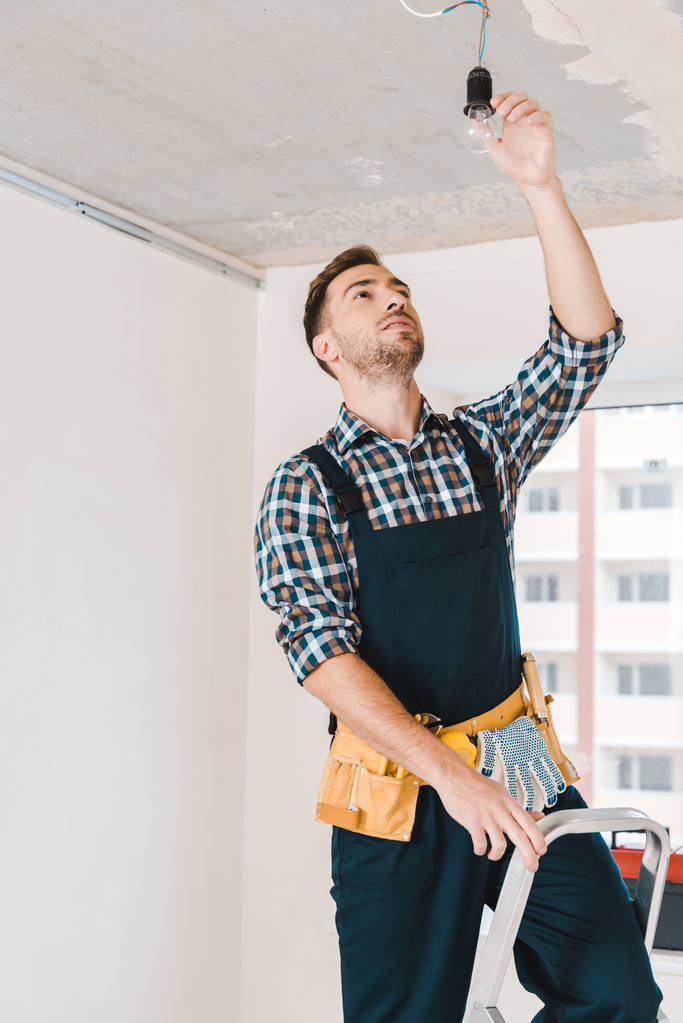 handsome handyman standing on ladder and looking at light bulb - Photo, Image