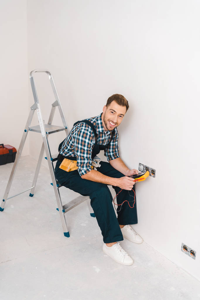 happy handyman holding digital multimeter and sitting on ladder near power socket  - Photo, Image