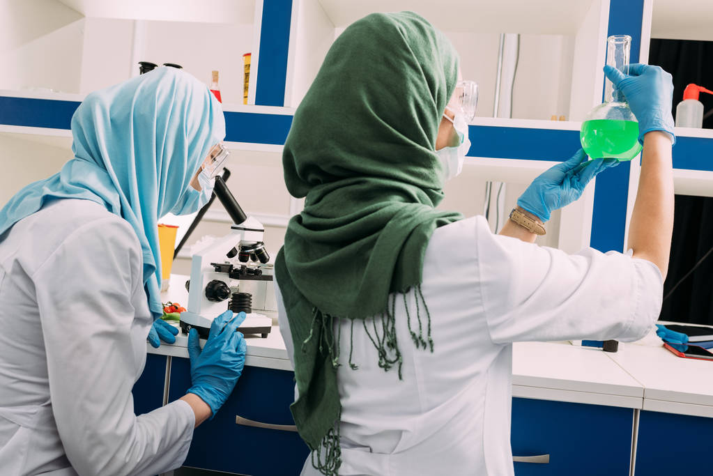 vista posterior de las científicas musulmanas que sostienen el frasco y usan el microscopio mientras experimentan en un laboratorio químico
 - Foto, Imagen