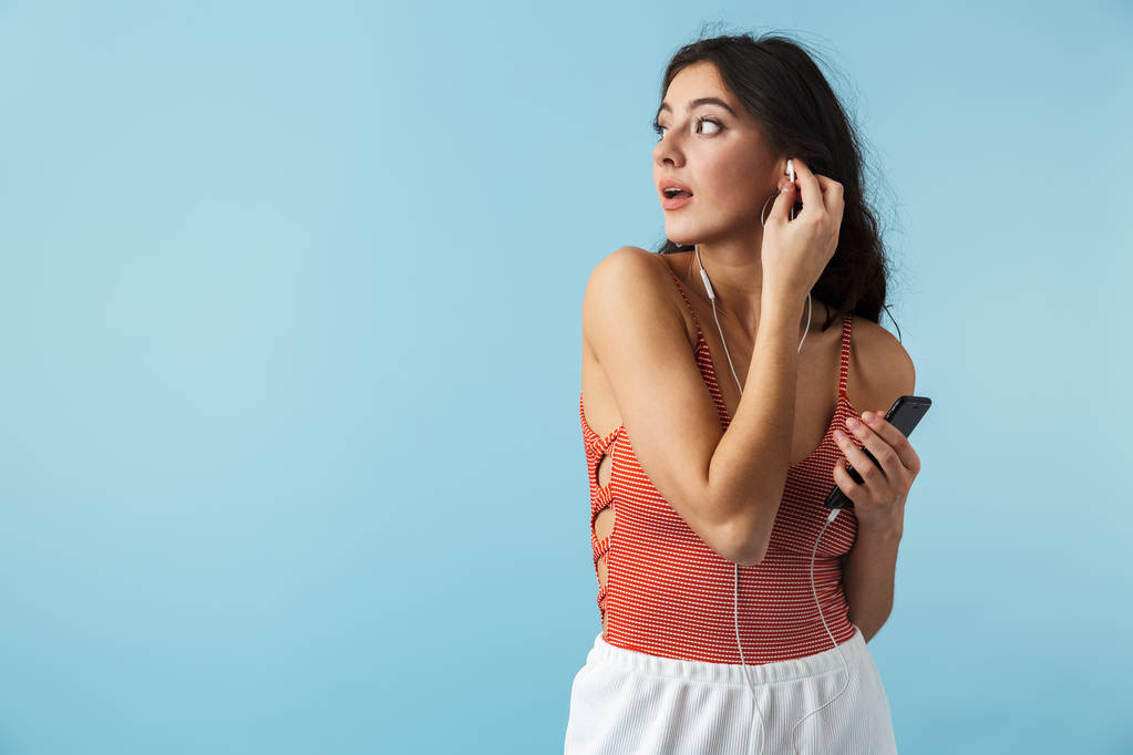 Прекрасна дівчина в літньому одязі стоїть ізольовано на синьому фоні, слухаючи музику з навушниками, тримаючи мобільний телефон
 - Фото, зображення