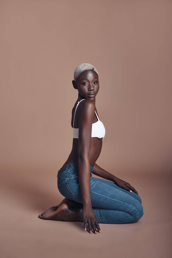 attraktive junge Afrikanerin sitzt vor braunem Hintergrund und trägt ein Oberteil mit Jeans  - Foto, Bild