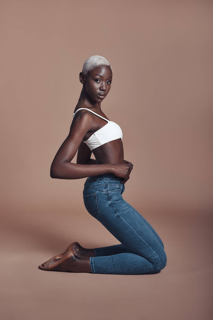 Aantrekkelijke jonge Afrikaanse vrouw zit tegen bruine achtergrond en dragen van boven met denim jeans  - Foto, afbeelding