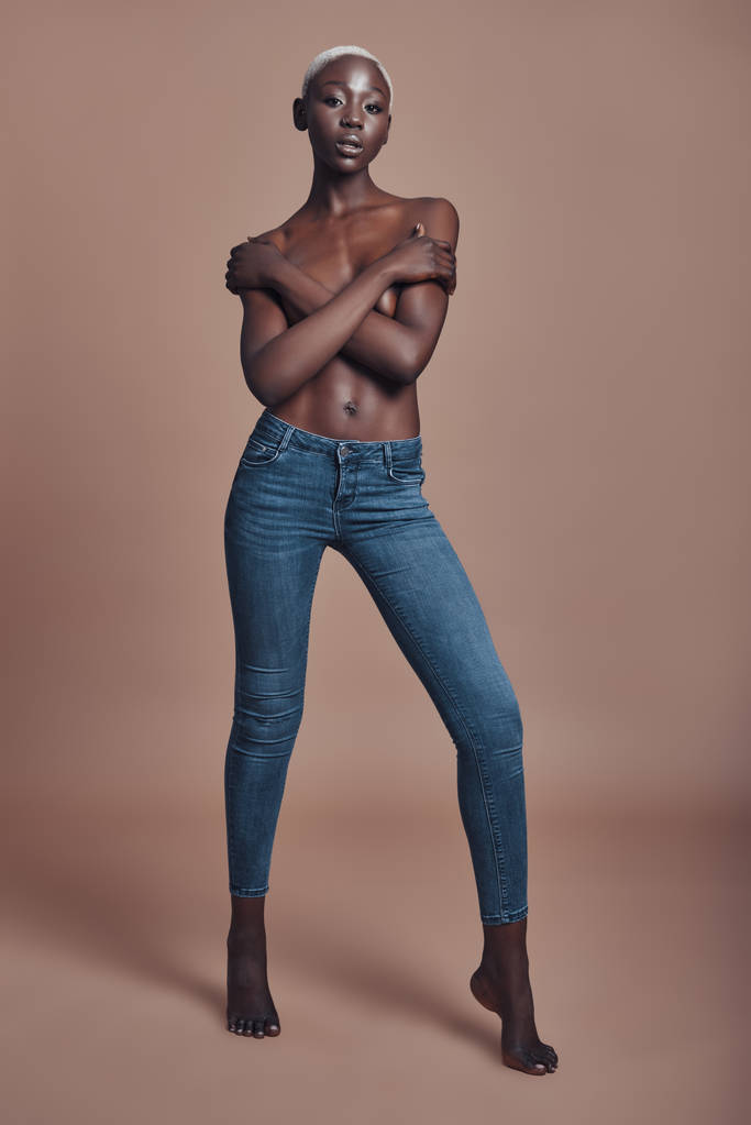 カメラ目線と茶色の背景に対する立ちながら手で胸を覆う魅力的な若いアフリカ女性の完全な長さ - 写真・画像