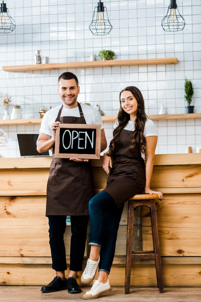 cashiers in aprons standing near wooden bar counter and holding chalkboard with open lettering in coffee house - Photo, Image