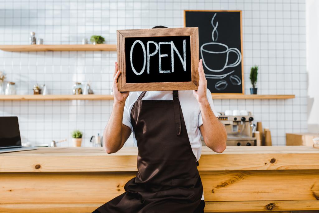 cashier in apron sitting on chair near wooden bar counter and holding chalkboard in front of face with open lettering in coffee house - Photo, Image
